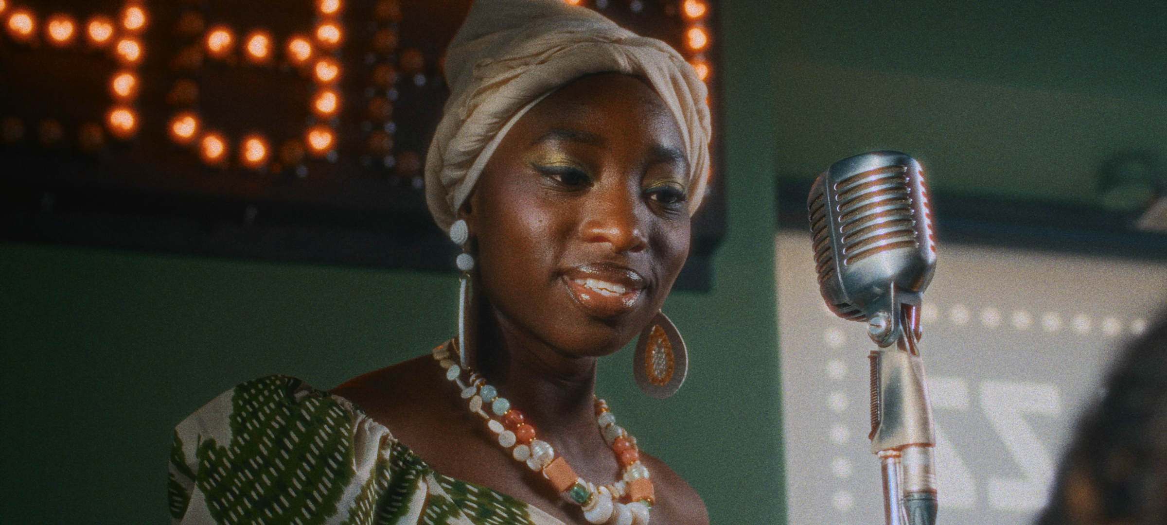 Bloom + Film Grain + Halation — Woman in a headwrap performing beside a vintage microphone on a softly lit stage, with bloom, film grain, and halation effects.