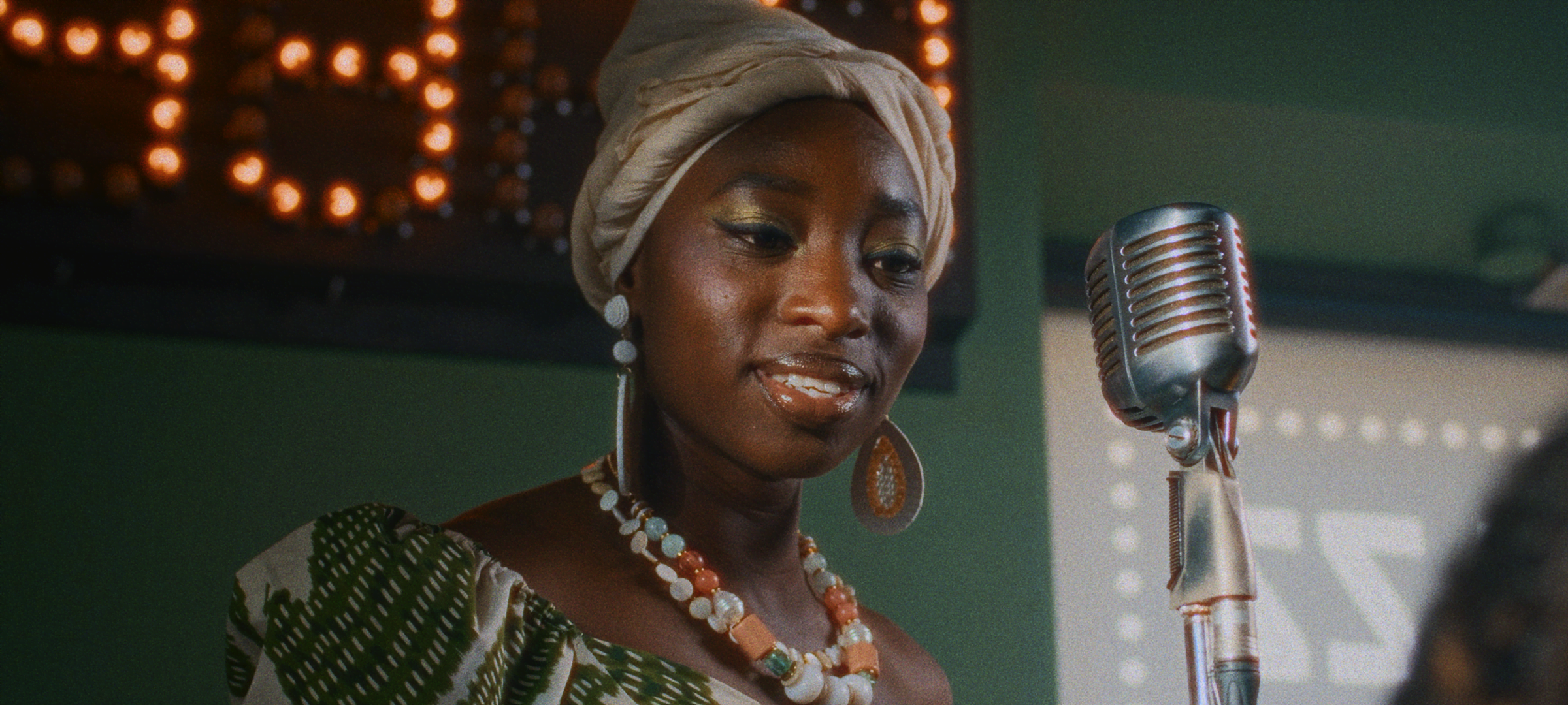Film Grain + Halation — Woman in a headwrap performing beside a vintage microphone on a softly lit stage, with film grain and halation effects.