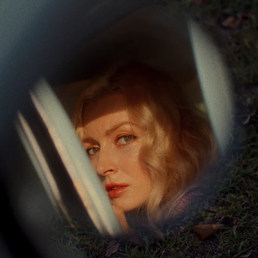 Kodak Kodachrome 64 Experimental — Blonde woman seen through a car window, framed by grass and soft reflections.