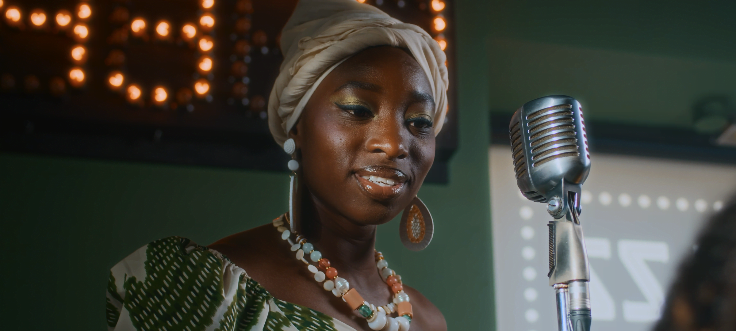 Bloom — Woman in a headwrap performing beside a vintage microphone on a softly lit stage, with bloom effect.