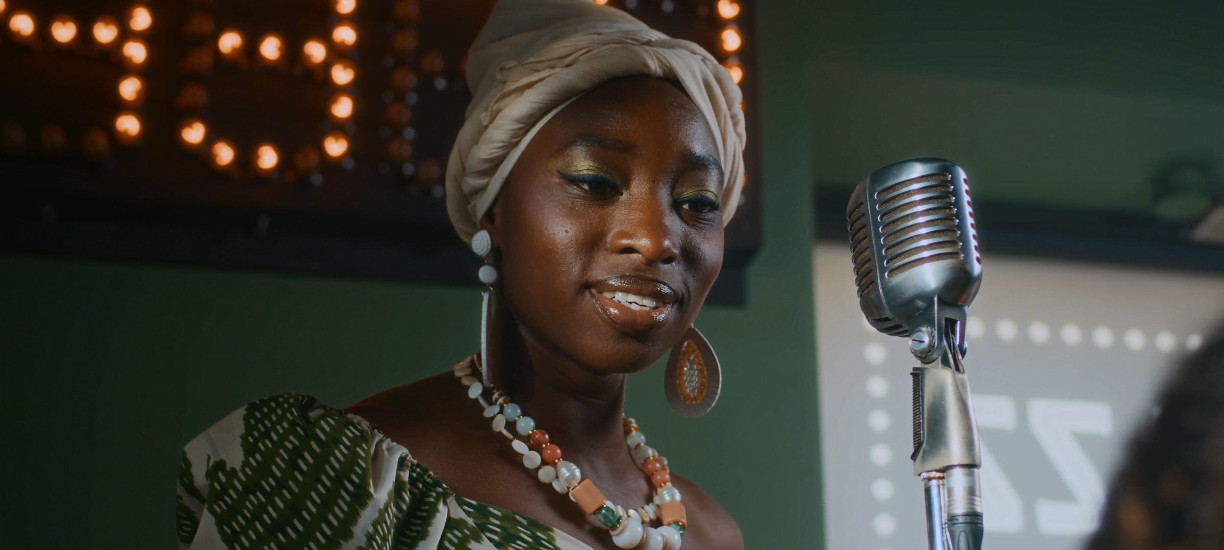 Woman in a headwrap performing beside a vintage microphone on a softly lit stage, with no added effects.