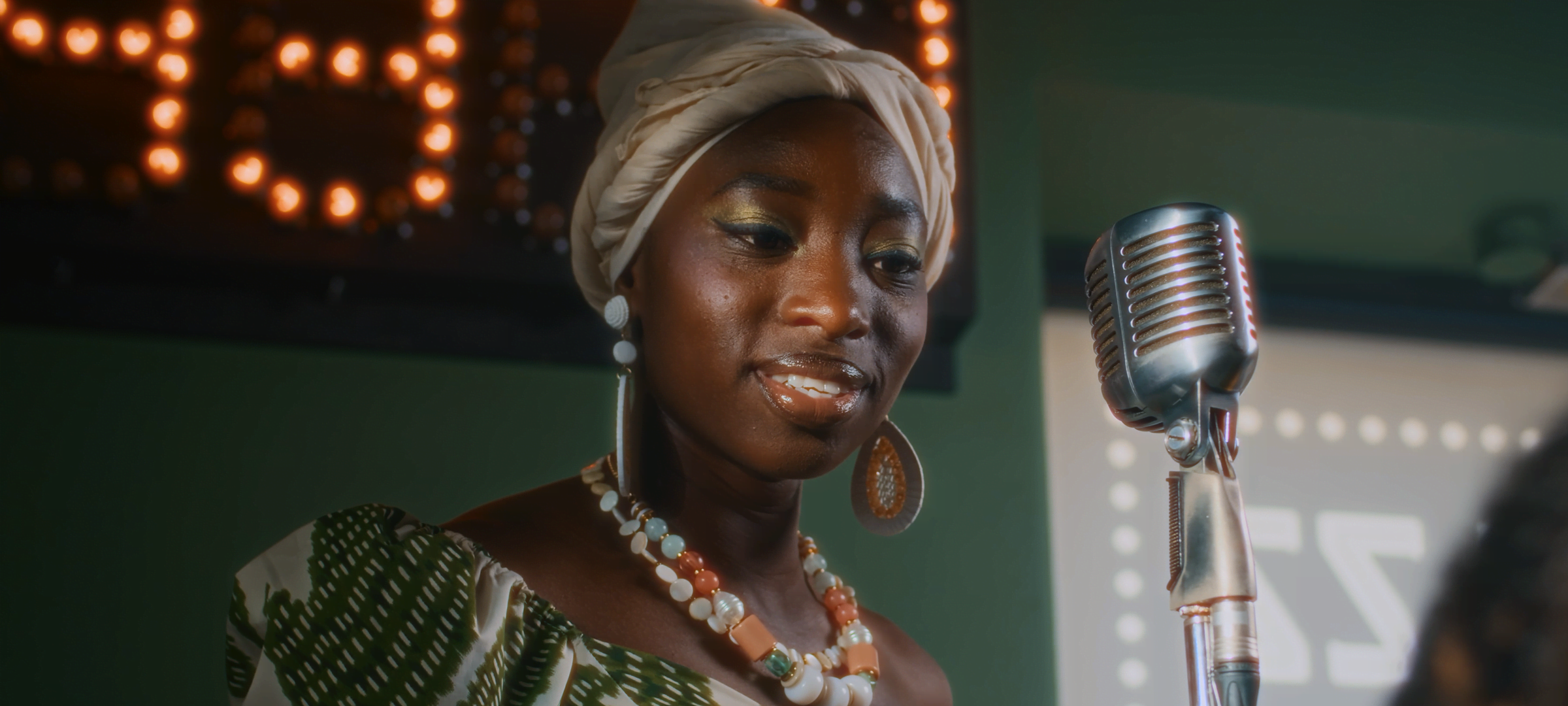 Bloom + Halation — Woman in a headwrap performing beside a vintage microphone on a softly lit stage, with bloom and halation effects.